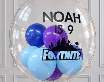 May include: A large, clear, spherical balloon containing smaller blue, light blue, and purple balloons. The exterior features black text reading NOAH IS 9 and a black Fortnite logo silhouette. A dark blue ribbon is tied at the base of the balloon.