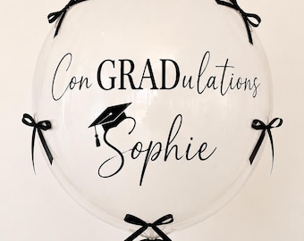 May include: A clear, round balloon featuring the black text "Con GRADulations" and the name "Sophie" in a cursive script, accompanied by a black graduation cap icon. The balloon is decorated with five small black ribbon bows attached to its surface.