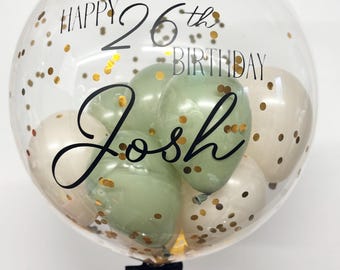 May include: A clear balloon filled with smaller balloons and gold confetti. The balloon has a black inscription that reads "Happy 26th Birthday Josh".