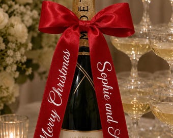 May include: A Moët & Chandon champagne bottle with a gold foil neck wrap and a red satin ribbon tied in a bow. The ribbon features white printed text that reads Merry Christmas on the left side and Sophie and Alan on the right side.