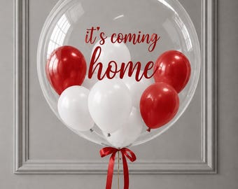 May include: A large, clear, spherical balloon containing smaller red and white balloons inside. The text 'it's coming home' is printed in red script on the surface of the clear balloon. A red ribbon is tied at the base of the balloon.