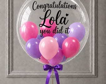 May include: A large clear bubble balloon containing smaller pink and purple balloons. The text "Congratulations Lola you did it" is printed in black on the surface of the clear balloon. A purple ribbon is tied in a bow at the base of the balloon.