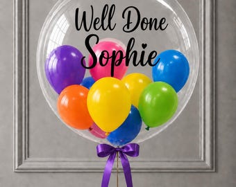 May include: A large clear bubble balloon containing smaller multi-colored balloons in purple, pink, blue, orange, yellow, and green. The text "Well Done Sophie" is printed in black script on the surface of the clear balloon, which is tied with a purple ribbon.
