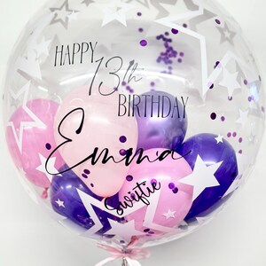 May include: A clear bubble balloon with purple and pink balloons inside. The balloon has white stars and purple confetti inside. The balloon has the text "Happy 13th Birthday Emma Swiftie" written on it.