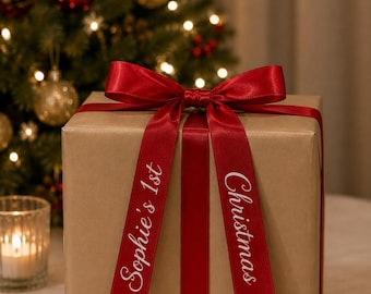 May include: A square gift box wrapped in plain brown paper, tied with a deep red satin ribbon featuring a bow on top. The ribbon tails are printed with the white text "Sophie's 1st" on the left and "Christmas" on the right side of the box.