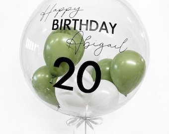 May include: A large clear balloon containing smaller olive green and white balloons. The exterior is printed with the black text Happy BIRTHDAY Abigail and the number 20. A white ribbon is tied at the base of the clear balloon.