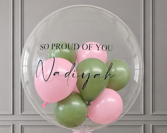 May include: A large, clear, spherical balloon containing smaller pink and olive green balloons. The text SO PROUD OF YOU is printed in black capital letters, and the name Nadiyah is written in a black cursive script on the clear surface.