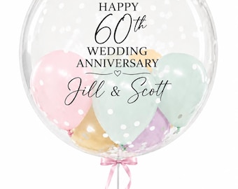 May include: A clear bubble balloon filled with smaller pastel-colored balloons in pink, mint green, yellow, and purple. The exterior features black text reading HAPPY 60th WEDDING ANNIVERSARY and the names Jill & Scott, finished with a pink ribbon.