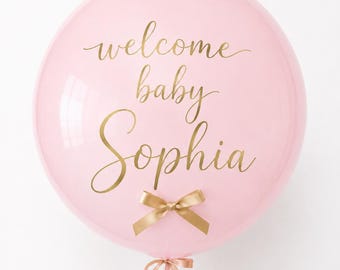 May include: A large, round, light pink balloon featuring the gold-colored text "welcome baby Sophia" in a cursive script. A small, gold-colored ribbon bow is attached to the center of the balloon, and a matching ribbon is tied at the base.