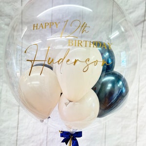 May include: A clear balloon filled with white and black balloons. The balloon has a gold inscription that reads "Happy 12th Birthday Hudson".