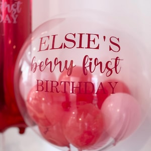 May include: A transparent balloon with the text "ELSIE'S berry first BIRTHDAY" in red. Inside are red and pink balloons. The balloon is tied with a red ribbon. A red balloon is visible in the background.
