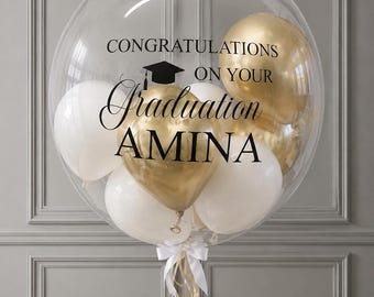 May include: A large clear bubble balloon filled with smaller white and metallic gold balloons. The exterior is printed with black text that reads CONGRATULATIONS ON YOUR Graduation AMINA, featuring a black graduation cap icon above the text.