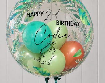 Könnte beinhalten: Ein klarer, runder Ballon, gefüllt mit kleineren Ballons in Blaugrün, Hellgrün und Orange. Der äußere Ballon ist mit einem grünen Aquarell-Blättermuster, dem Text "HAPPY 2nd BIRTHDAY" und einer schwarzen Strichzeichnung eines Monsters bedruckt.