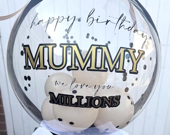 May include: A clear spherical balloon filled with smaller cream-colored balloons and black confetti. The exterior features black and gold text reading "happy birthday MUMMY we love you MILLIONS" and a black bow. It sits on a black base marked with "30".