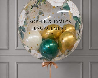 May include: A large clear balloon filled with smaller gold, white, and dark green balloons. The exterior features a floral print of white roses and green leaves, with the black text "SOPHIE & JAMIE'S ENGAGEMENT" printed in the center. A thin ribbon is tied at the base.