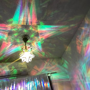 以下が含まれることがあります： 星型のガラス製のランプシェードが天井に虹色の光のパターンを投影しています。ランプシェードは黒いコードから吊り下げられており、内部の電球によって照らされています。