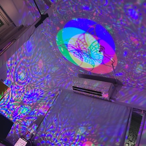 Puede incluir: Un proyector proyecta un patrón colorido de mariposas y flores en una pared y un techo. El proyector es visible en la esquina superior derecha de la imagen.