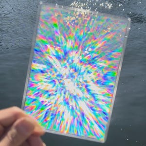 以下が含まれることがあります： 虹色の光沢のある抽象的なデザインが施された透明なプラスチックカード。デザインは光の爆発や流れ星に似ています。