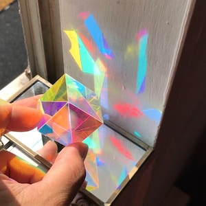 May include: A clear glass prism cube refracting light, casting rainbow colours on a white surface.