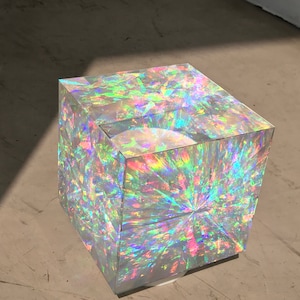 Puede incluir: Un cubo transparente con colores arcoíris iridiscentes. El cubo está hecho de vidrio y tiene una superficie reflectante.