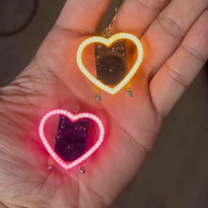 以下が含まれることがあります： 手の中に、ピンクと黄色の2つのハート型のLEDペンダントがあります。各ペンダントは回路基板の裏地と、吊り下げ用の金色のループが付いています。ペンダントは小さく、ジュエリーやクラフトプロジェクトに適しています。