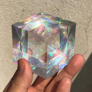 Puede incluir: Un cubo transparente iridiscente con colores arcoíris que se reflejan en la superficie. El cubo está hecho de vidrio o cristal y tiene un acabado liso y pulido.