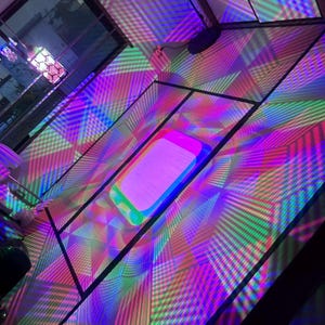 Pode incluir: Um dispositivo projetor de luz retangular colocado no chão projeta um padrão geométrico vibrante e multicolorido de linhas e grades cruzadas em tons de magenta, ciano, verde e amarelo sobre as superfícies e o chão ao redor.