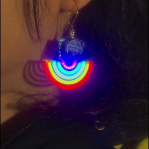 Puede incluir: Un par de pendientes LED de colores arcoíris con una pequeña placa de circuito adjunta. Los pendientes están iluminados con una variedad de colores, incluyendo rojo, naranja, amarillo, verde, azul, índigo y violeta. La placa de circuito tiene el texto "We encourage oddity" impreso en ella.