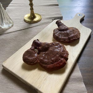May include: Two dark brown, mushroom-shaped objects rest on a light-colored wooden cutting board. The board has a handle and is placed on a beige tablecloth. A gold-colored candle holder is in the background.