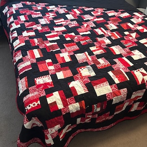 May include: A handmade quilt with a patchwork design in red, white, and black. The quilt features a repeating pattern of squares and rectangles, with a black border and backing. The quilt is displayed on a bed.