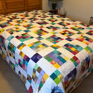 May include: A colorful patchwork quilt with a white background and a variety of colors, including purple, green, yellow, and blue. The quilt is made of squares of different fabrics and is quilted with a simple pattern. The quilt is on a bed with a wooden headboard.
