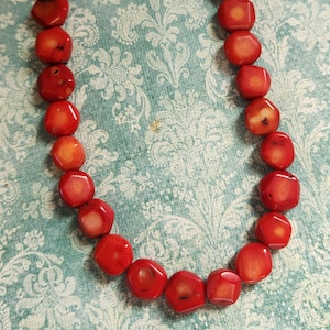 May include: A vibrant red coral necklace. The necklace features polished, irregularly shaped coral beads strung together with a silver-toned chain. The beads are a rich, warm red color, and the necklace is displayed against a patterned background.