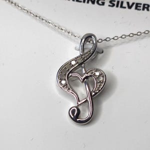 May include: Sterling silver necklace with a treble clef pendant. The pendant is embellished with small diamonds and features a heart-shaped design. The necklace is on a white background, with the text "1/10 CTW DIAMONDS".