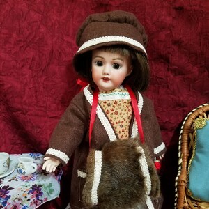 May include: A vintage doll wearing a brown coat, hat, and fur purse. The doll has a floral print dress and white shoes. A small table with teacups and a floral tablecloth is in the background.