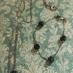 K&ouml;nnte beinhalten: Ein silberfarbenes Halsketten- und Armband-Set. Die Halskette hat eine lange Kette mit einem gro&szlig;en, dunkelblauen, kristallbedeckten Kugelanh&auml;nger. Das Armband hat &auml;hnliche Kristallkugeln. Beide Teile werden auf einem gemusterten Hintergrund pr&auml;sentiert.