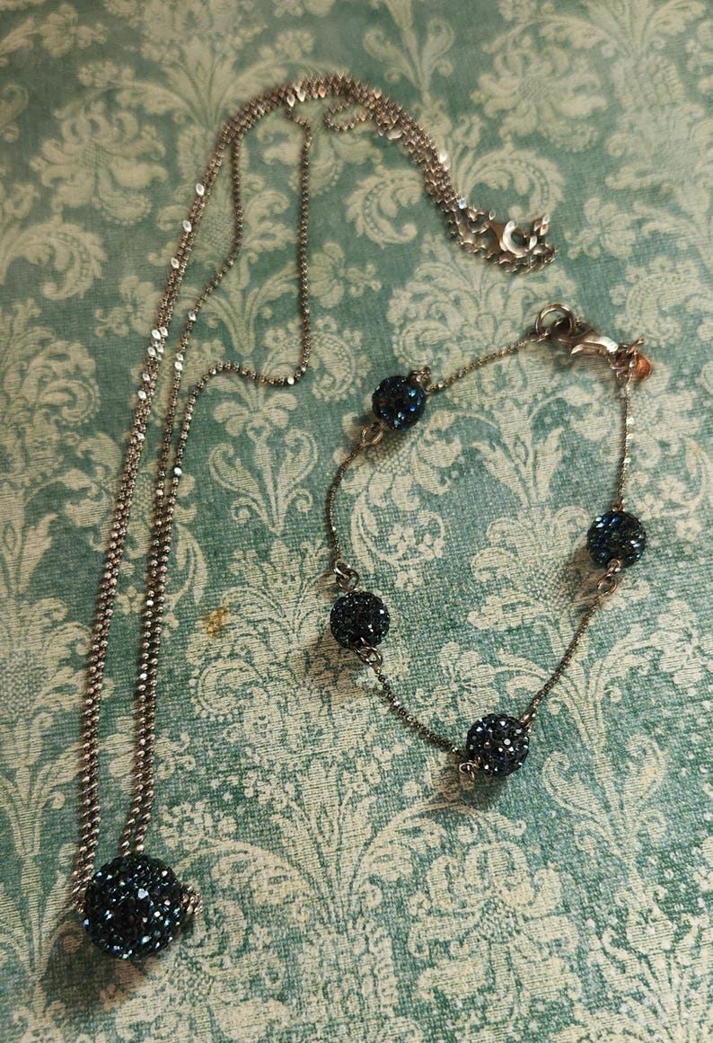 K&ouml;nnte beinhalten: Ein silberfarbenes Halsketten- und Armband-Set. Die Halskette hat eine lange Kette mit einem gro&szlig;en, dunkelblauen, kristallbedeckten Kugelanh&auml;nger. Das Armband hat &auml;hnliche Kristallkugeln. Beide Teile werden auf einem gemusterten Hintergrund pr&auml;sentiert.