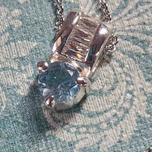 May include: A silver necklace with a light blue gemstone pendant. The pendant is set in a silver frame with smaller clear gemstones.