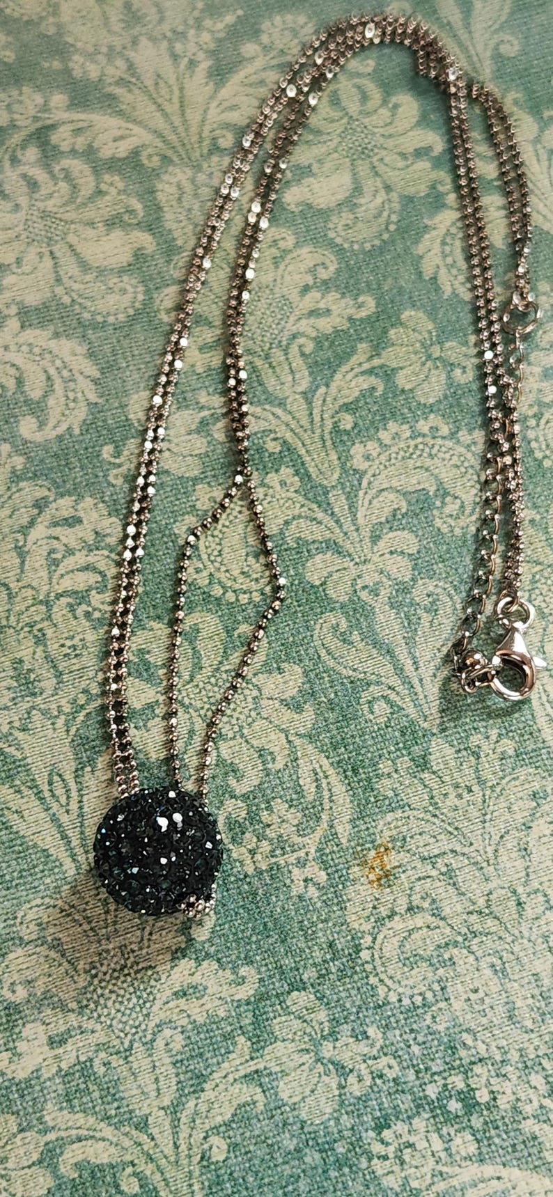 K&ouml;nnte beinhalten: Silberfarbene Halskette mit einem runden, schwarzen Anh&auml;nger, der mit glitzernden Kristallen besetzt ist. Die Kette besteht aus kleinen, reflektierenden Perlen. Die Halskette liegt auf einer t&uuml;rkisfarbenen Oberfl&auml;che mit Muster.