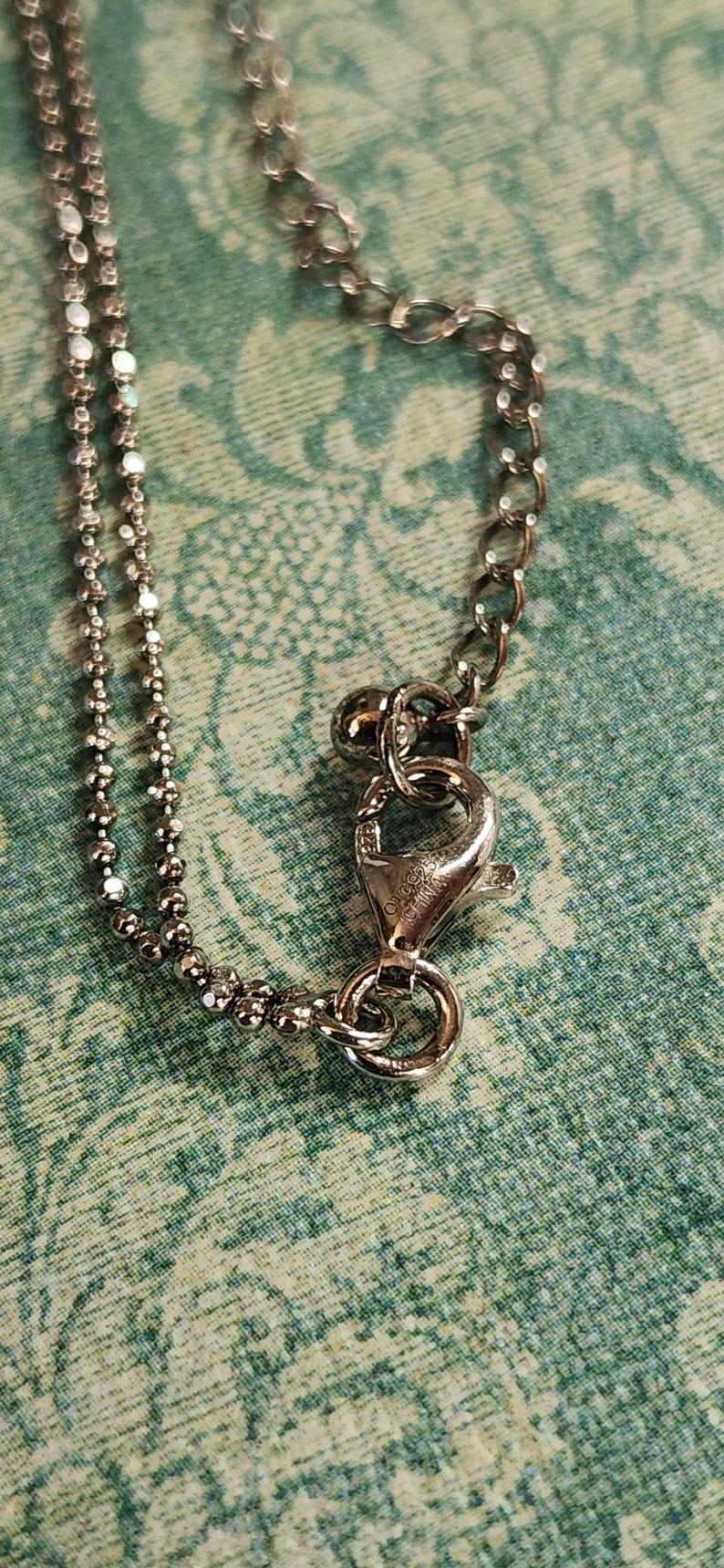 K&ouml;nnte beinhalten: Silberfarbene Halskette mit Karabinerverschluss. Die Kette hat auf einer Seite ein Kugelkettendesign und auf der anderen Seite eine traditionellere Kette. Der Verschluss ist an einer Kette mit gr&ouml;&szlig;eren Gliedern befestigt. Der Hintergrund ist ein gr&uuml;n-wei&szlig; gemusterter Stoff.