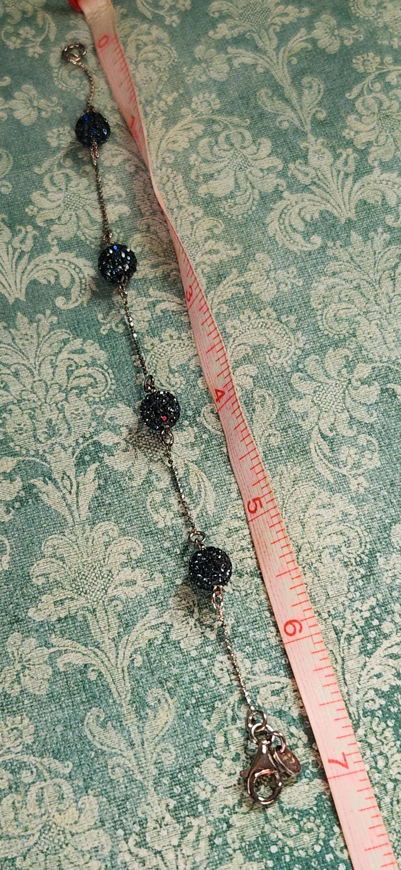 K&ouml;nnte beinhalten: Eine silberne Halskette mit dunkelblauen, kristallbedeckten Perlen. Ein rosa Ma&szlig;band liegt neben der Halskette und zeigt Messungen in Zentimetern an. Der Hintergrund ist ein t&uuml;rkisfarbenes Blumenmuster.