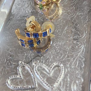 May include: Three pairs of gold hoop earrings with different colored gemstones. The first pair has a cluster of pastel pink, blue, and green gemstones. The second pair has a row of blue square gemstones. The third pair is a heart shape with clear gemstones.
