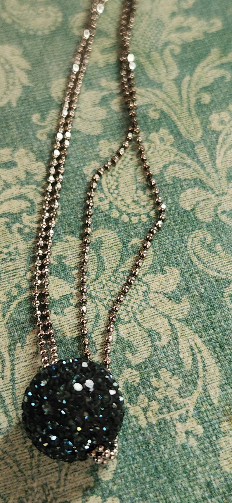 K&ouml;nnte beinhalten: Halskette mit einem dunklen, runden Anh&auml;nger, der mit glitzernden schwarzen und irisierenden Kristallen bedeckt ist. Die Kette besteht aus kleinen, verbundenen Perlen in Bronzefarbe, vor einem gr&uuml;nen floralen Hintergrund.