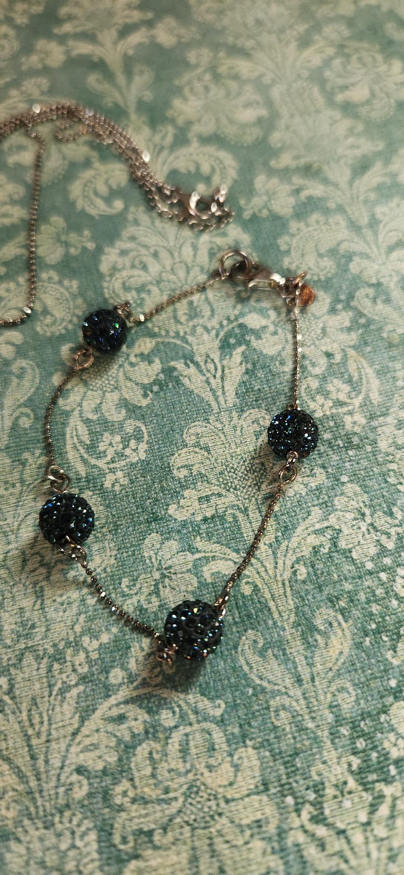 K&ouml;nnte beinhalten: Ein silbernes Armband mit vier dunkelblauen, kristallbedeckten Perlen. Eine passende Halskette ist ebenfalls zu sehen. Der Schmuck befindet sich auf einem teal-farbenen, gemusterten Hintergrund.