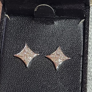 May include: A pair of rose gold stud earrings in a black jewelry box. Each earring is a four-pointed star shape, with a central cluster of small, clear stones. The earrings are displayed on a textured black surface.