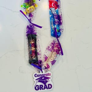 Puede incluir: Un collar de caramelos con varios caramelos envueltos en celofán transparente, atados con cinta morada. El collar incluye una etiqueta que dice "Congrats GRAD" en morado y blanco.