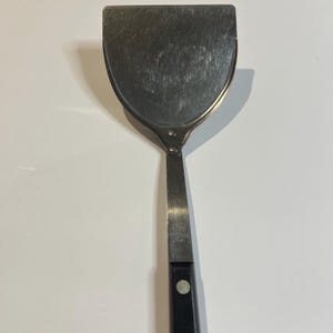 May include: A stainless steel spatula with a wide, rounded head and a long, narrow handle. The handle is black with a single silver rivet. The spatula is designed for cooking and food preparation.