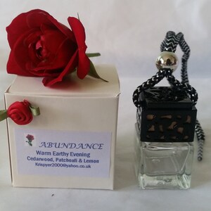 May include: A white box with a red rose on top. The box has a label that says "ABUNDANCE Warm Earthy Evening Cedarwood, Patchouli & Lemon Krispyer2006@yahoo.co.uk". Next to the box is a small glass bottle with a black metal cage and a black and white string hanging from it.