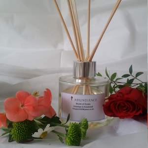 May include: A clear glass bottle with a silver lid and wooden sticks, filled with a light yellow liquid. The bottle has a label that reads "Abundance World of Roses Geranium & Rosewood Krissper2000@yahoo.co.uk". The bottle is surrounded by pink and red flowers.