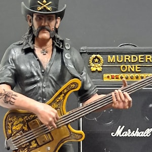 Op de afbeelding: Een zwart en gouden beeldje van een muzikant die basgitaar speelt. Het beeldje staat op een ronde basis met het woord "Motorhead" erop. De muzikant draagt een zwart leren jack, een zwarte broek en zwarte laarzen. De basgitaar heeft een gouden afwerking en een zwarte slagplaat. Het beeldje staat voor twee zwarte Marshall-versterkers met de tekst "Murder One" op de bovenste versterker.