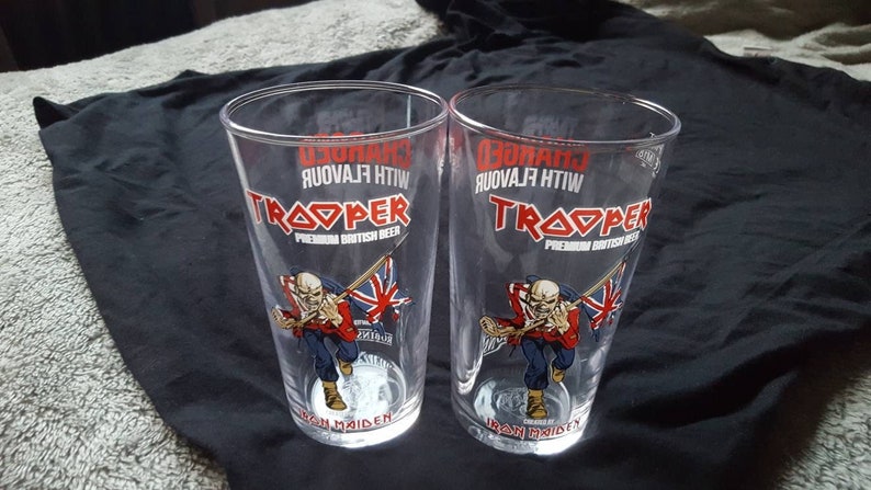 Puede incluir: Dos vasos de pinta de vidrio transparente con un gr&aacute;fico rojo, blanco y azul de un hombre sosteniendo una bandera. El texto en el vaso dice "Trooper Premium British Beer" e "Iron Maiden".